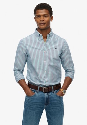 Chemise à col boutonné avec des rayures verticales bleues et blanches ; col, manches longues, logo subtil ; associée à un jean bleu et une ceinture marron.
