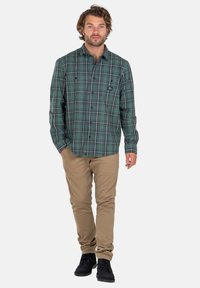 Mann mit lockigem Haar, der ein grünkariertes Hemd mit Knöpfen, beige Hosen und schwarze Schuhe trägt und mit einer Hand in der Tasche dasteht.