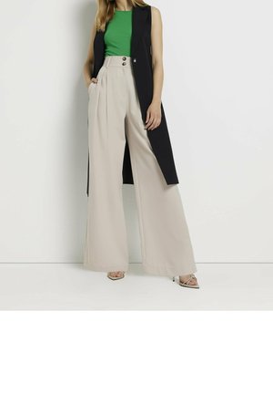 Frau trägt grünes ärmelloses Oberteil, lange schwarze Weste, hoch taillierte weite beige Hose und Riemchensandaletten mit hohen Absätzen, steht vor weißer Wand.