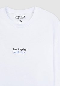 T-shirt bianca oversize, con scollatura rotonda. La grafica frontale in nero e blu riporta la scritta "Los Angeles Unhaltable Creators."