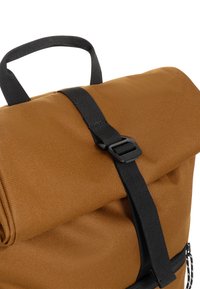 Mochila de tela marrón con parte superior enrollada, asegurada con una correa negra y detalle de hebilla. Superficie texturizada, equipada con un asa superior.