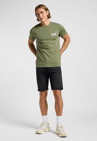 Oliwkowa bawełniana koszulka z białym logo "Lee", w połączeniu z ciemnoniebieskimi szortami dżinsowymi. Model ma na sobie kremowe sneakersy i białe skarpetki.