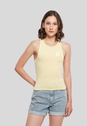 Jeune femme portant un haut sans manches jaune pâle avec un petit motif de marguerite blanche et un short en jean bleu clair roulé sur un fond gris.