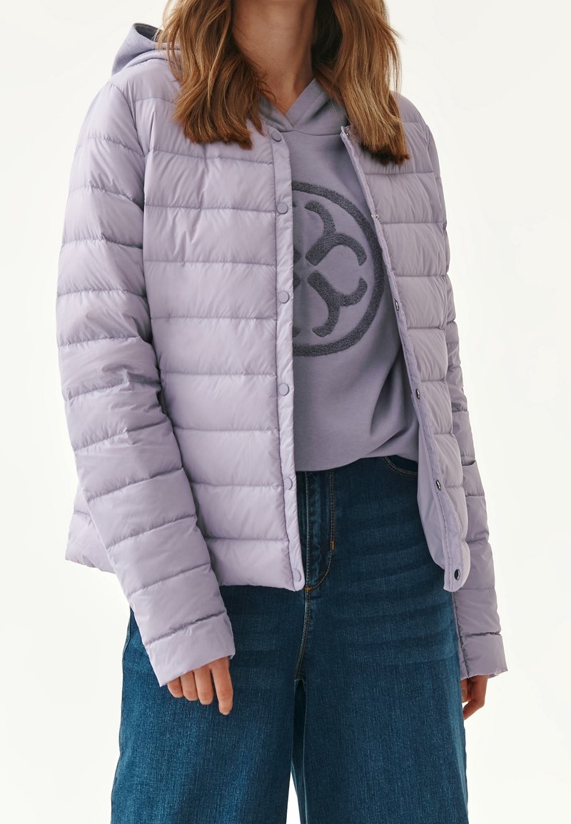 Lyse lilla, fyldige jakke med horisontale quiltede mønster og trykknapper, lagdelt over en grå hoodie med et rundet logo, parret med blå jeans.