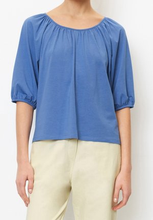Vrouw draagt een loszittende blauwe top met een geplooide hals en driekwart pofmouwen, gecombineerd met lichtbeige broek.