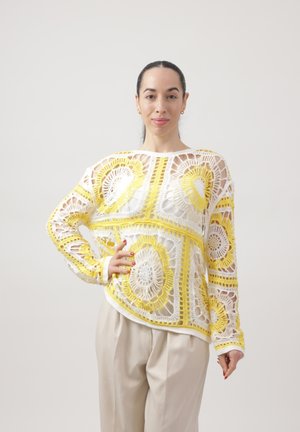 Vrouw met donker haar in een witte en gele gehaakte trui en beige broek, staand met één hand op haar heup tegen een effen achtergrond.