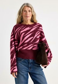 Maglione lavorato a maglia con un audace motivo astratto rosa e bordeaux, maniche ampie e orlo a costine; abbinato a una borsa in suede marrone scuro.