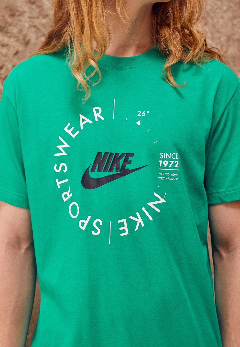 Vihreä puuvillainen T-paita, jossa on musta Nike-logo ja valkoinen teksti "SPORTSWEAR" ja "SINCE 1972", sekä graafisia suunnitteluelementtejä.