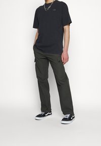 Person klädd i en svart The North Face-t-shirt, mörkgröna cargobyxor, svartvita sneakers och ett silverhalsband, stående med händerna i fickorna.