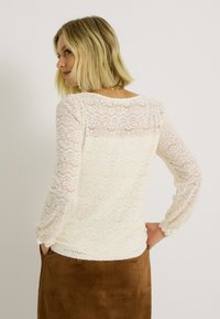 Haut en dentelle blanc cassé avec des manches longues et un motif ondulé et texturé. Présente un empiècement légèrement transparent et des poignets élastiqués.