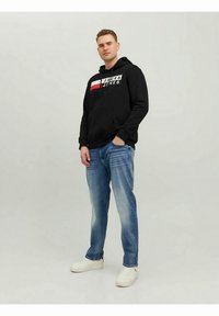 Crna majica s kapuljačom s bijelim i crvenim detaljem na logotipu, u kombinaciji sa svijetloplavim trapericama i bijelim tenisicama, stojeći ispred neutralne pozadine.