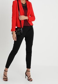 Blazer rouge cintré en tissu lisse, associé à un haut foncé et un pantalon noir fuselé. Accessoires incluant un petit sac à main noir et des talons à fines lanières.