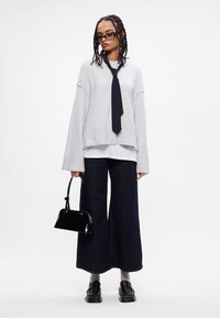 Ανοιχτό γκρι φαρδύ πουλόβερ με μπλε γραβάτα, συνδυασμένο με σκούρα παντελόνια μποξέρ. Κρατά μια γυαλιστερή μαύρη τσάντα, φορώντας μαύρα παπούτσια και γκρι κάλτσες.