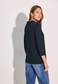 Musta, pitkähihainen paita, jossa on kaulus, pyöristetty helma ja hienovarainen tekstuuri. Yhdistetty vaaleansinihin, rentoon farkkuihin.