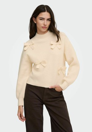 Femme portant un pull crème côtelé avec quatre nœuds en tissu, associé à un pantalon marron foncé, posant avec la main dans la poche devant un fond uni.