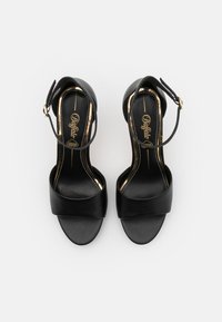 Μαύρες δερμάτινες ψηλοτάκουνες σάντες με φαρδύ μπροστινό λουράκι, λουράκι στον αστράγαλο με χρυσό κούμπωμα και λεία υφή. Χρυσές λεπτομέρειες στην εσωτερική σόλα.