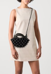 Frau in einem ärmellosen beigefarbenen Karokleid, die eine kleine schwarze Handtasche mit Netzdesign und Kettenriemen hält.