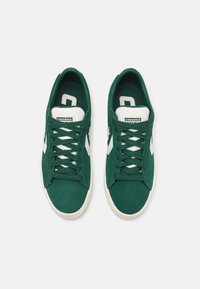 Adidași din suede verde cu accente și șireturi albe, având o talpă plată din cauciuc și o suprafață texturată. Logo-ul Converse pe limbă.