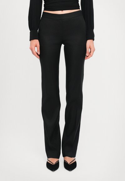 Schwarze maßgeschneiderte Hose mit glatter Textur, slim fit und geradem Bein, ausgestattet mit einem mittelhohen Bund. Kombiniert mit spitzen Absatzschuhen.