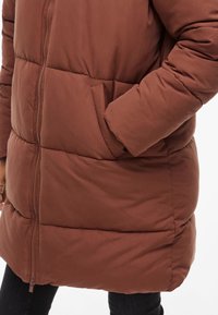 Cappotto imbottito marrone con zip e tasca laterale, caratterizzato da pannellature cucite per texture e calore. Tessuto liscio con una leggera lucentezza.