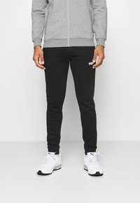 Pantaloni sportivi neri aderenti con un logo bianco sulla coscia, abbinati a scarpe da ginnastica bianche e un maglione grigio con zip.