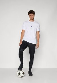 Biele športové tričko so zeleno-fialovým logom, v kombinácii s čiernymi teplákmi s bielymi bočnými pruhmi, obutými do čiernych futbalových kopačiek, na lopte.