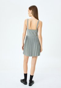 Robe plissée grise avec un dos carré, des bretelles larges et un tissu texturé. Portée avec des chaussettes noires et des chaussures noires brillantes.
