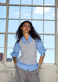 Jeune femme aux longs cheveux bouclés et lunettes, portant une chemise bleue, un gilet gris et un pantalon gris, se tient avec confiance près d'une grande fenêtre.