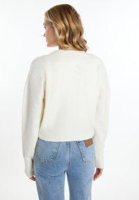 Kremafarbener, verkürzter Pullover mit geripptem Saum und weiten Ärmeln, kombiniert mit hellblauen, hochgeschnittenen Jeans, die ein braunes Leder-Patch auf der Rückseite haben.