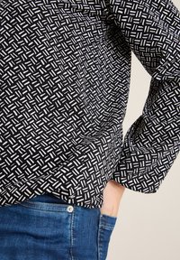 Top nero con motivo a fantasia bianca e design geometrico, maniche lunghe, tessuto morbido. Abbinato a jeans blu con tasca frontale e bottone argentato.