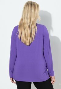 Blondýna zády, oblečená v dlouhém fialovém topu a černých kalhotách, stojící před jednoduchým bílým pozadím.