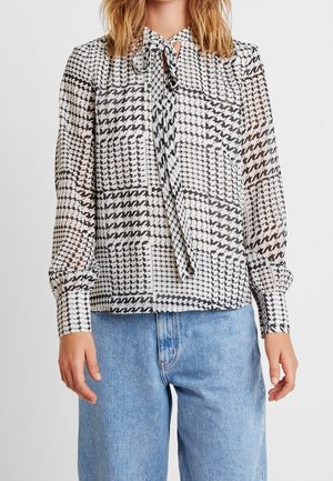 Vrouw draagt een zwart-wit patchwork pied-de-poule blouse met een nekstrik en lange mouwen, gecombineerd met lichtblauwe spijkerbroek.