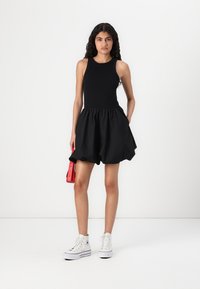 Zwarte mouwloze jurk met een aansluitende top en een uitlopende rok. Gecombineerd met witte hoge sneakers en een kleine rode handtas.