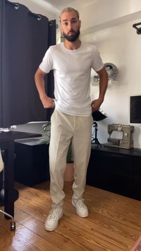T-shirt bianco a girocollo, pantaloni larghi color crema e scarpe da ginnastica bianche. Presenta un pavimento in legno e tende scure in un contesto di stanza.