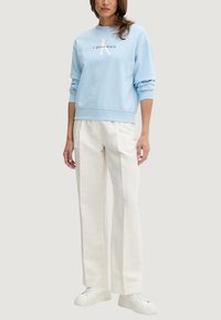 Felpa azzurro chiaro con logo "Calvin Klein" bianco, abbinata a pantaloni larghi color crema e sneaker bianche, che mostra un outfit casual.