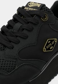 Μαύρα αθλητικά παπούτσια με λείο πάνω μέρος και υφασμάτινες λεπτομέρειες, με μαύρα κορδόνια, χρυσή πινακίδα με λογότυπο και υφασμένη σόλα.