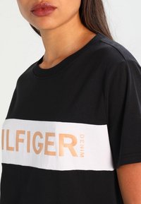Černé bavlněné tričko s bílým vodorovným pruhem a nápisem "TOMMY HILFIGER" ve světle oranžové barvě. Kulatý výstřih, bezseamové ramenní švy.
