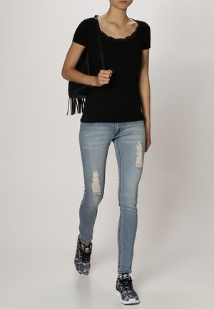 Kvinna klädd i svart kortärmad topp, slitna ljusblå skinny jeans, mönstrade svarta sneakers och bär en svart fransväska på axeln.