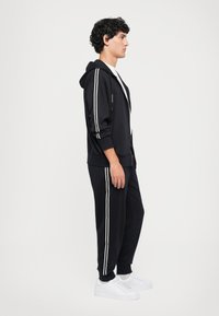 Svart joggedress med hvite sidestroiper, med en glidelås-hettegenser og matchende slim-passform bukser. Brukt med hvite sneakers, glatt tekstur.