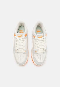 Zapatillas deportivas en blanco y gris con acentos en naranja. Hechas de materiales lisos y texturizados. Cuentan con un diseño de cordones y una zona de los dedos perforada.