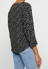 Camisetta nera con motivo floreale bianco, maniche 3/4 e vestibilità rilassata. Tessuto morbido e leggero. Indossata con jeans azzurri chiari.