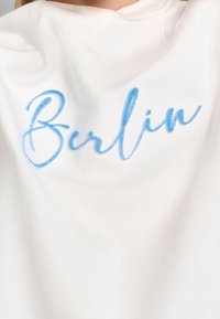 Felpa bianca con testo ricamato in blu che dice "Berlino". Il tessuto appare morbido con una superficie liscia e un collo rotondo.