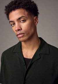 Camicia a maniche corte di un verde scuro con collo e bottoni. Indossata sopra una maglietta nera, con il tessuto testurizzato visibile.