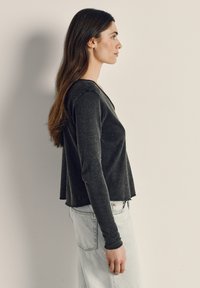 Pitkähiuksinen nainen seisoo sivuprofiilissa, yllään tummanharmaa pitkähihaisen paita ja vaaleansiniset farkut, taustalla beige seinä.