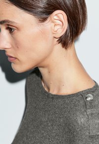 Szara dzianinowa sweterka z okrągłym dekoltem, krótkimi rękawami oraz ozdobnym srebrnym guzikiem po lewej stronie na ramieniu.