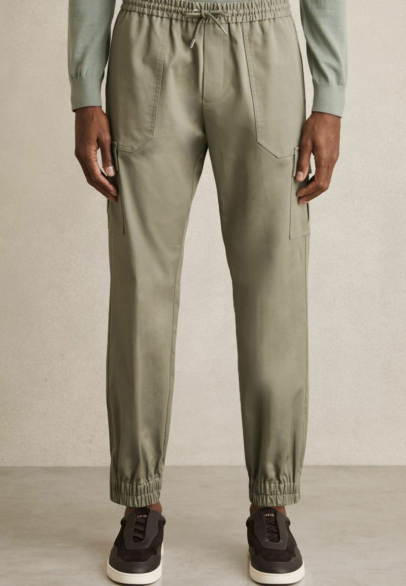 Pantalones cargo en color caqui, tapered, con cinturilla elástica y puños en los tobillos. Presentan dos bolsillos frontales y un bolsillo lateral. Tejido suave al tacto.