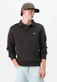 Tamsiai rudas polo tipo megztinis su raukinėtomis rankovėmis, su žaliu Lacoste logotipu. Suderintas su šviesiai mėlynomis džinsinėmis kelnėmis ir raštuota kepure.