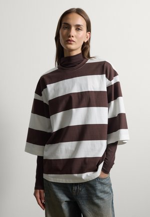 Frau mit glatten braunen Haaren trägt ein lockeres braun-weiß gestreiftes Kurzarmtop über einem braunen Langarmshirt und blauen Jeans.