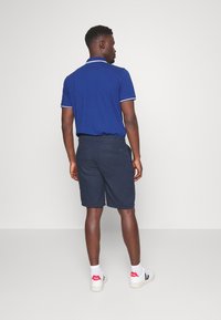 Mørkeblå polo med hvid trim, parret med mørke shorts og hvidt fodtøj med røde accenter, set bagfra.