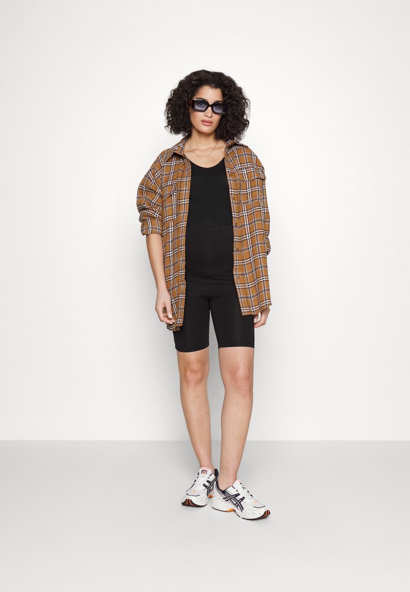 Un débardeur noir et un short de vélo associés à une chemise à carreaux brune surdimensionnée. Accessoirisé avec des lunettes de soleil foncées et des baskets blanches.